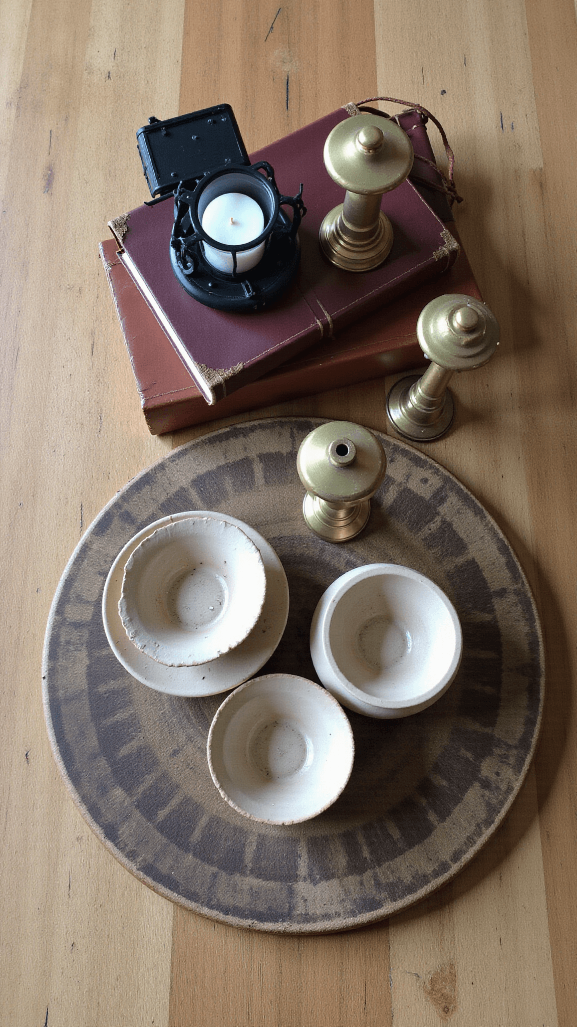Overhead view of a vintage-styled coffee table with artisanal pottery on a bread board, a stack of leather-bound books with brass candlesticks, and a black metal lantern on a weathered oak surface