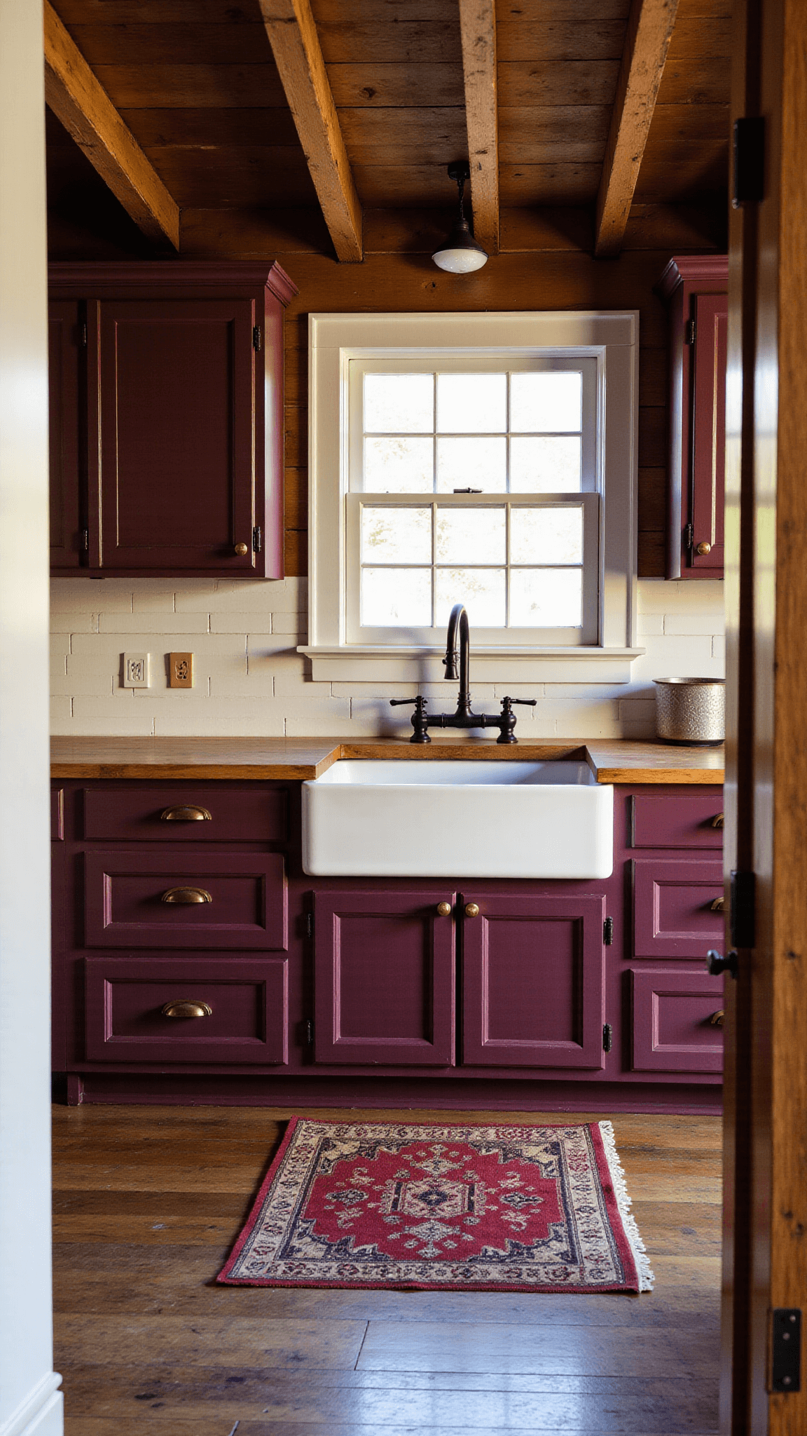 Late afternoon light illuminating a rustic 14'x18' farmhouse kitchen with exposed wooden beams, distressed burgundy cabinets with vintage brass cup pulls, warm butcher block countertops, white shiplap backsplash, weathered antique rug runner and black cast iron hardware.