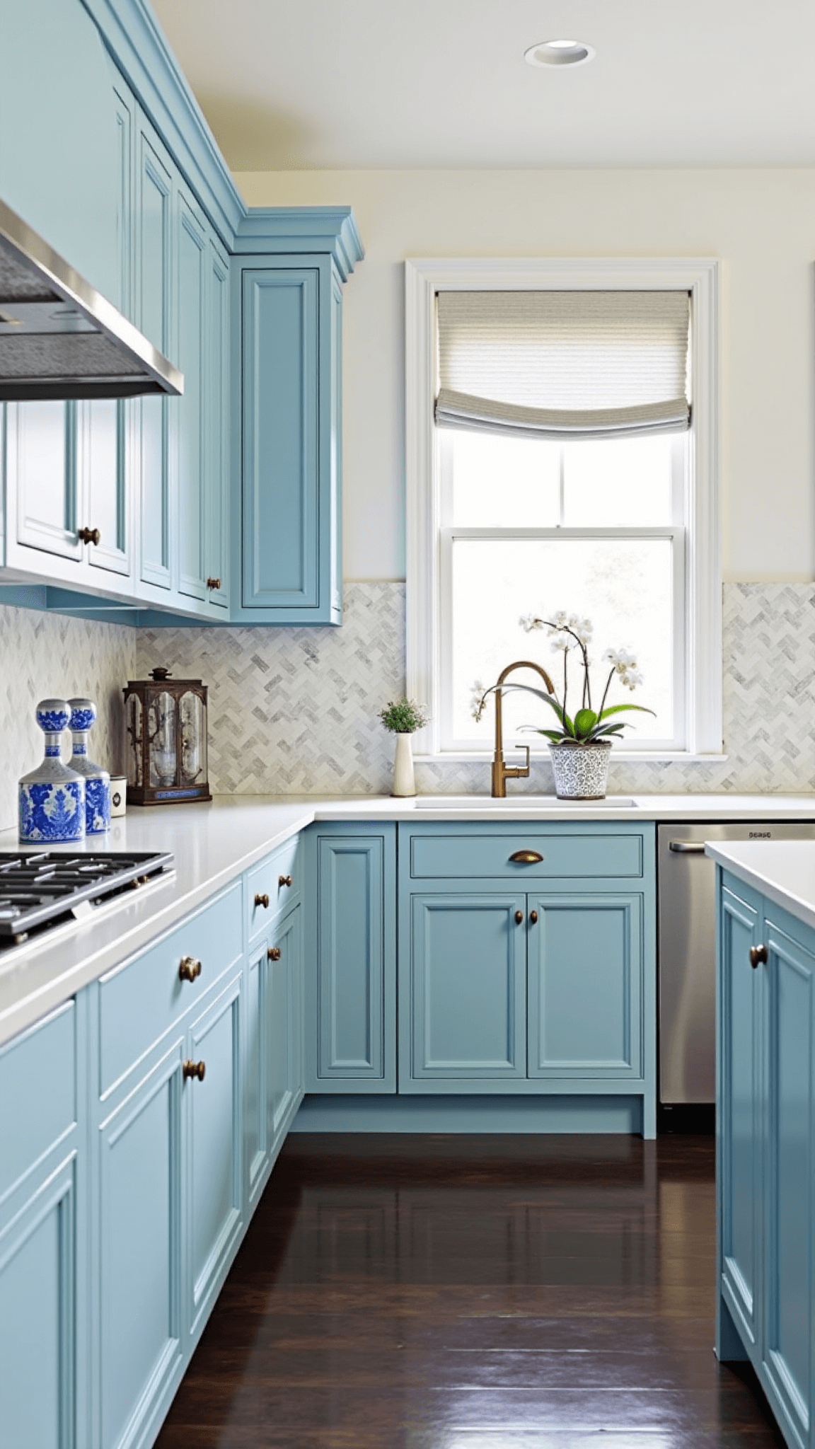 Transitional kitchen with powder blue inset cabinets, marble herringbone backsplash, and gray oak floors decorated with chinoiserie, crystal decanters, and potted orchids in the mid-morning light.
