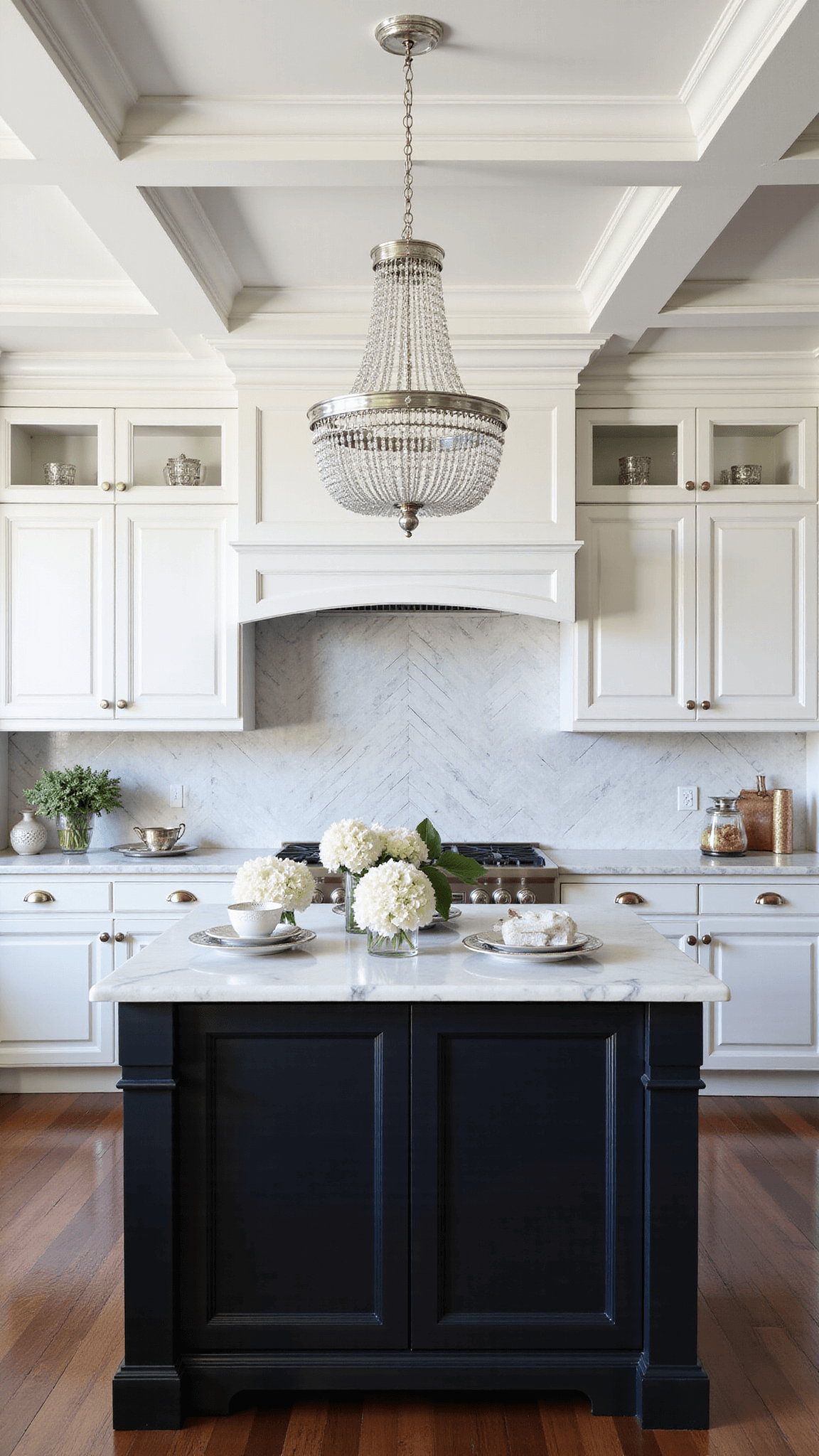 Elegant traditional kitchen with white cabinets, black glazed island, marble counters, herringbone backsplash, and crystal chandelier, styled with silver serving pieces and white hydrangeas, with soft afternoon light casting a timeless mood.
