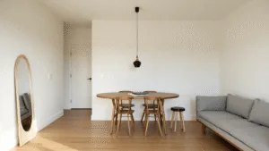 "Minimalist design studio apartment with white walls, oak floors, grey sofa and round walnut table, bathed in warm golden sunlight reflected by an oversized mirror."